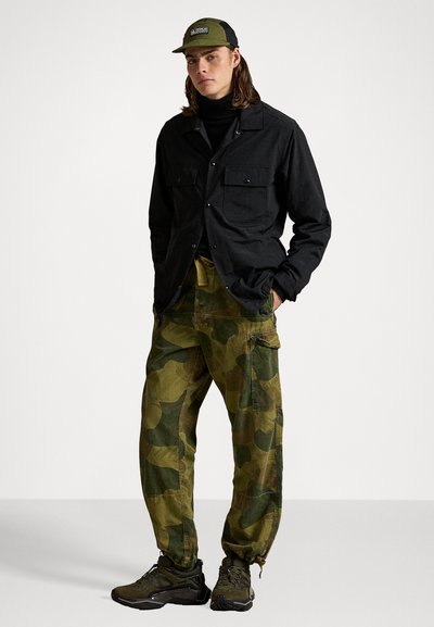 Cămașă neagră cu nasturi, pantaloni cargo verzi cu camuflaj, adidași negri și o șapcă de baseball verde. Design casual și funcțional, cu buzunare.