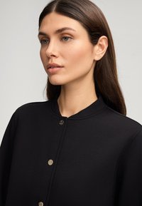 Veste noire avec un col rond, fermeture à pression et texture lisse. Présente des coutures discrètes et un design minimaliste.
