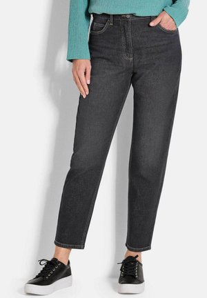 Persona che indossa jeans neri slim-fit, sneakers nere con suola bianca e una maglia a maniche lunghe texturizzata in teal, in piedi contro un muro bianco.