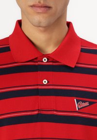 Polo rayé rouge et marine avec col, deux boutons et un logo en patch sur le côté gauche. Texture en coton, présentant des rayures horizontales.