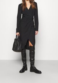 Schwarzes Wickelkleid mit langen Ärmeln, V-Ausschnitt und seitlichem Schlitz. Kombiniert mit kniehohen schwarzen Lederstiefeln und einer strukturierten schwarzen Handtasche.