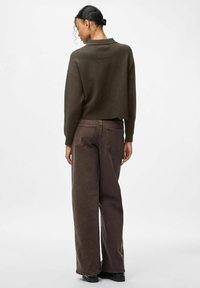 Kvinna med lockigt hår i en mörk knut bär en mörkbrun kort tröja och bruna jeans med vida ben, stående mot en enkel ljus bakgrund.