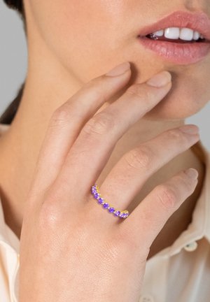 Anello d'oro con gemme viola disposte in fila, indossato su una mano. La mano ha un tono di pelle naturale e tocca delicatamente il viso.