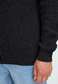 Maglione nero lavorato a maglia con un motivo testurizzato, polsini e orlo a coste. Indossato sopra un jeans azzurro chiaro, che mette in mostra una vestibilità aderente.
