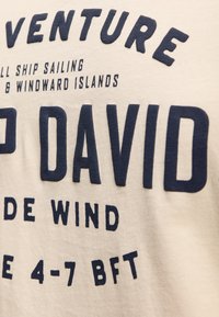 Beige cotton t-shirt featuring bold navy blue text. Text includes "ADVENTURE," "CAP DAVID," and nautical references. Soft texture.