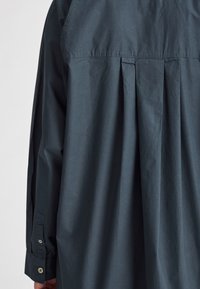 Tmavě modrá košile na knoflíky s nařaseným zadním dílem a dlouhými rukávy. Vyrobena z hladkého materiálu s volným střihem a knoflíkovými manžetami.