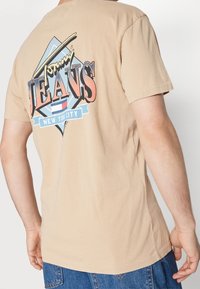 Světle hnědé bavlněné tričko s velkým vícebarevným grafickým potiskem "Tommy Jeans" ve tvaru diamantu, nošené s modrými džínovými šortkami.