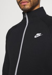 Muž v černé Nike mikině na zip s bílým zipem a logem, viditelnou dolní částí obličeje a krku.