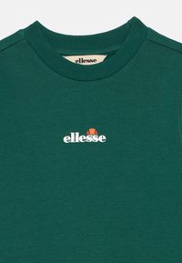 Zaļa sporta krekls, kas izgatavots no mīksta auduma, ar ribotu apkakli. Uz tā ir redzams baltais logo "ellesse" ar nelielu sarkanu grafiku virs tā.