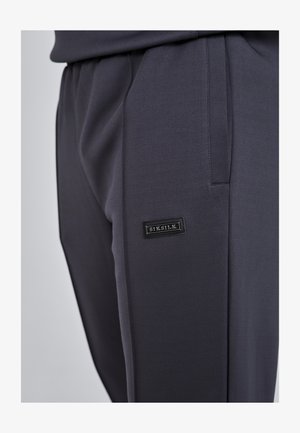 Dunkelgraue Jogginghosen aus weichem Stoff, mit Seitentaschen und einem schwarzen Markenlabel, das auf dem linken Oberschenkel aufgestickt ist. Glatte Textur.