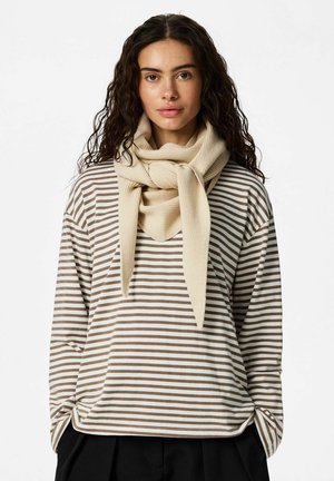 Junge Frau mit lockigem Haar, die einen beigen Schal und ein braun-weiß gestreiftes langärmeliges Shirt trägt und vor einem einfarbigen Hintergrund steht.