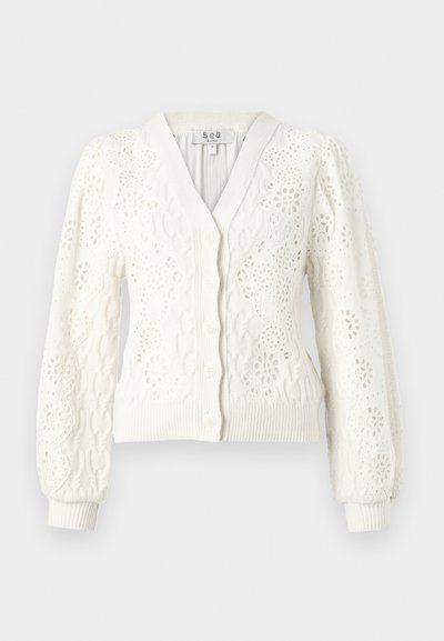 Balts adīts cimdiņš ar V veida kakla izgriezumu, pogu aizdari un smalkām acu detaļām. Puffētas piedurknes un ribota apakšmala pievieno struktūru.