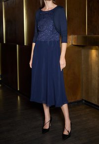 Mujer con un vestido de encaje azul marino y un cárdigan corto, usando tacones negros de punta, de pie sobre un suelo de madera oscura junto a paneles de pared de bronce.