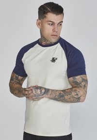 T-Shirt mit kurzen Ärmeln, cremefarbener Körper und marineblauen Raglanärmeln. Mit kleinem schwarzem Logo auf der linken Brust und einer taillierten Passform. Glatte Textur.