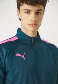 Puma fél cipzáras dzseki sötét teal színben, rózsaszín vállbetétekkel. A dzseki textúrált gallérral és egy nagy rózsaszín logóval rendelkezik a mellkason.