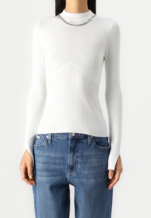 Vrouw draagt een getailleerde witte geribbelde trui met duimgaten in de mouwen en blauwe spijkerbroek, geaccentueerd met een zilveren ketting.
