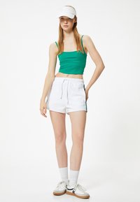 Top corto verde con tirantes finos, combinado con pantalones cortos blancos que presentan una franja lateral. Una visera blanca y calcetines deportivos completan el look.