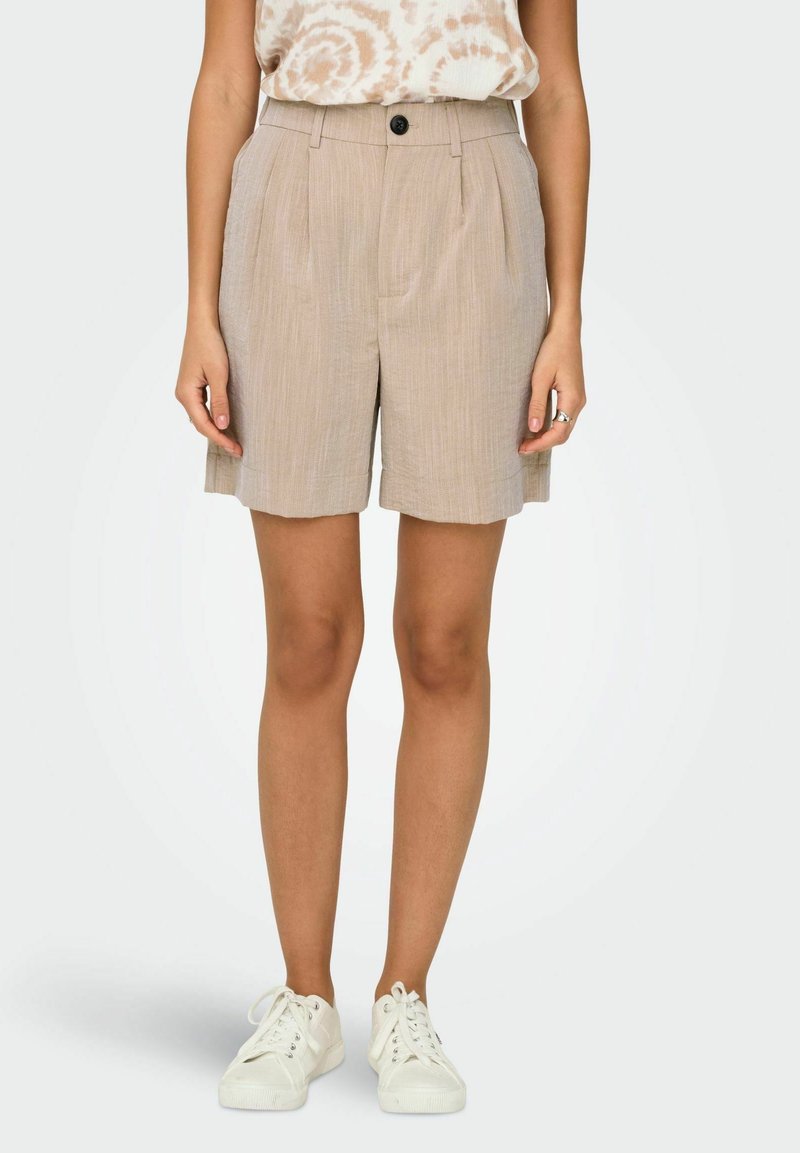 Femme portant un short plissé beige taille haute avec un bouton et des baskets blanches, debout devant un fond clair uni.