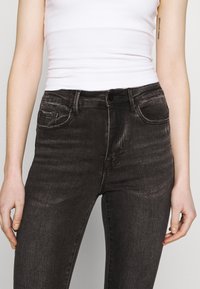 Persona vestida con jeans ajustados de cintura alta en negro desgastado y una camiseta sin mangas blanca, con las manos relajadas a los lados, frente a un fondo simple.