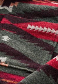 Tissu en laine avec un design géométrique comportant des couleurs rouge, bordeaux et gris. Surface texturée avec une sensation douce et épaisse. Motifs zigzag et triangle distincts.