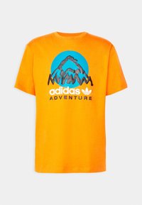 Oranžové bavlněné tričko s kruhovým motivem s horami a nápisem „adidas ADVENTURE“ v černé a bílé barvě.