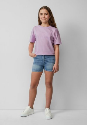 Jasnofioletowa koszulka z krótkim rękawem zestawiona z niebieskimi dżinsowymi szortami i białymi sneakersami. Ubranie charakteryzuje się swobodnym krojem i prostym wzornictwem.