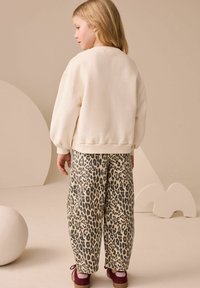 Hanorac bej cu mâneci bufante asortat cu pantaloni de trening cu imprimeu leopard. Fundalul prezintă forme abstracte în crem. Pantofi roșu-maro vizibili.