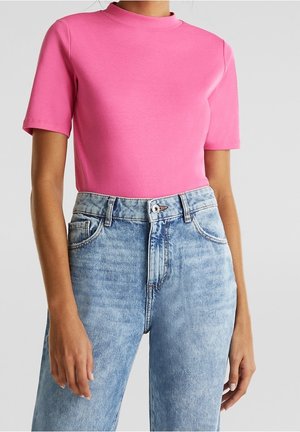 Frau trägt ein knallpinkes, kurzärmeliges, figurbetontes Oberteil, das in hoch taillierte, hellblaue Jeans gesteckt ist, vor neutralem Hintergrund.