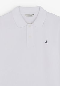 Polo blanc en coton, avec un col classique, une patte de boutonnage à trois boutons et un petit logo de tête de mort et os croisés en bleu marine sur la poitrine gauche.