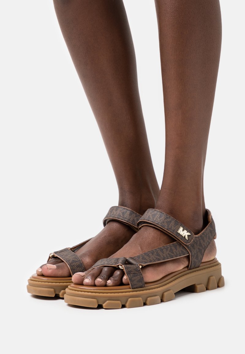 Sandales marron foncé à motifs avec semelles épaisses couleur sable portées pieds nus sur une peau foncée, avec un logo MK doré sur la bride de cheville.