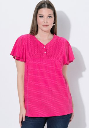 Kvinde iført en lyserød kortærmet bluse med små hvide prikker, plisseret foran og to knapper, kombineret med mørkeblå jeans.