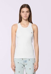 Una giovane donna con capelli castani lisci indossa una canotta bianca attillata e jeans azzurri chiari decorati con borchie riflettenti argento.