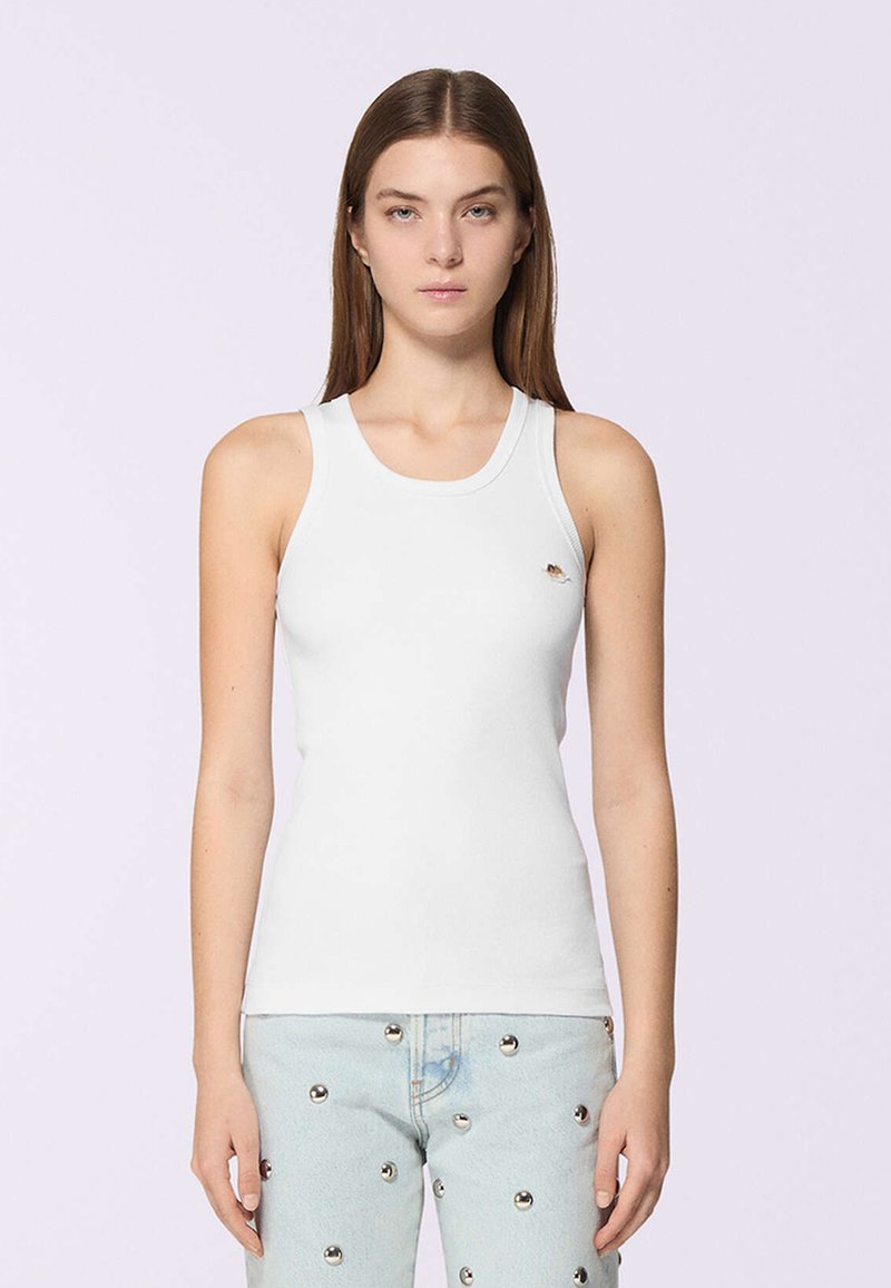 Una giovane donna con capelli castani lisci indossa una canotta bianca attillata e jeans azzurri chiari decorati con borchie riflettenti argento.