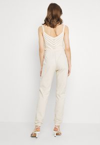 Beige jumpsuit met een gestructureerde V-hals bodice en elastische taille. Heeft aansluitende manchetten en ontspannen broekspijpen. Sleehakken in het wit.