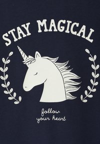 Marineblaues Hemd mit einem weißen Einhorn-Graphic, darüber der Text "BLEIB MAGISCH" und darunter "folge deinem Herzen", akzentuiert mit Blattdesigns.