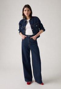 Jachetă din denim albastru închis și blugi cu talie înaltă asortați cu pantofi roșii cu toc. Jacheta are un design clasic, cu buzunare frontale și nasturi.