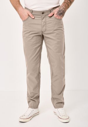 Mann trägt hellbraune Freizeithosen, weiße Turnschuhe und ein weißes Hemd, Hände in den Taschen, mit sichtbar tätowiertem Unterarm.