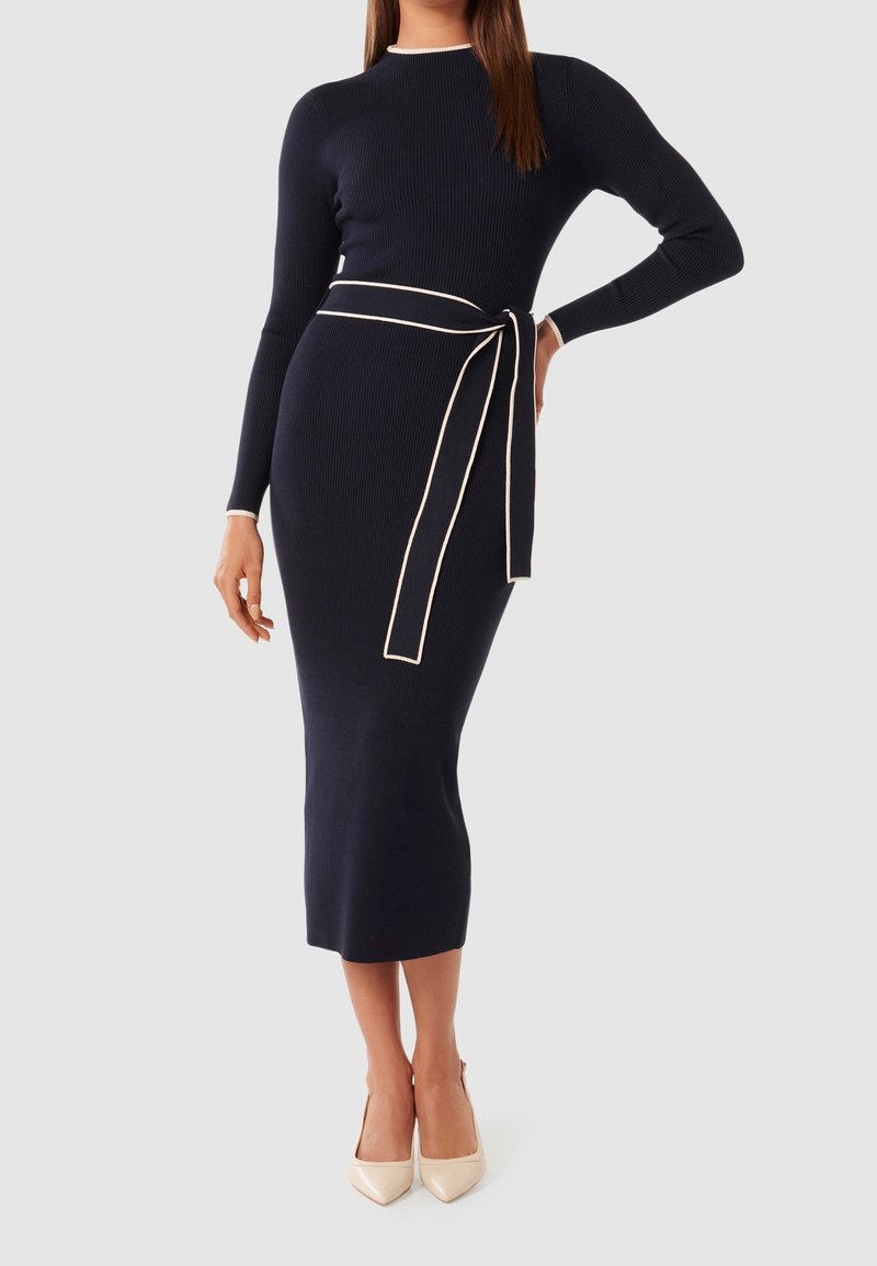 Femme portant une robe en maille côtelée bleu marine ajustée avec des manches longues, des bordures blanches, une ceinture nouée à la taille et des talons pointus beiges.
