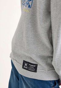 Sudadera gris con estampado gráfico azul, puños y dobladillo acanalados. Presenta una etiqueta negra con texto blanco en la parte inferior izquierda.