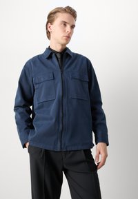 Tumesinine tõmblukujope, mis on valmistatud puuvillast. Sellel on kaks rinna taskut ja kaelus, koos mustade pükstega. Sile tekstuur.