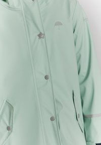 Ljust grön regnjacka i vattentåligt material. Har silverknappar, en dekorativ paraply-logotyp och en sidficka.