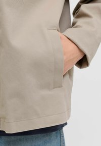 Main glissée dans la poche latérale d'un manteau beige porté sur un jean bleu, sur un fond gris uni.