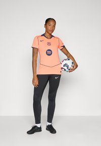 Lichtoranje voetbalshirt met marineblauwe accenten en logo, gecombineerd met zwarte strakke leggings met textuurpatronen. De speler houdt een multicolore voetbal vast.
