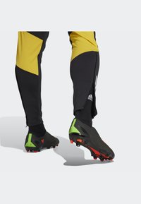 Schwarze Fußballschuhe mit grünen und roten Akzenten, strukturierten Oberflächen, getragen mit schwarzen Socken in Knöchelhöhe und gelb akzentuierter Hose mit Reißverschlüssen.