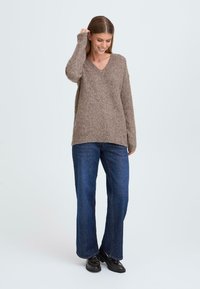Brown, textured, V-neck sweater with long sleeves, paired with blue wide-leg jeans and black shoes. Minimalist design against a light background.