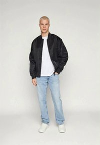 Čierna bomber bunda, biele tričko, svetlo modré džínsy a biele tenisky. Model stojí na jednotnom pozadí, predvádzajúc detaily oblečenia.