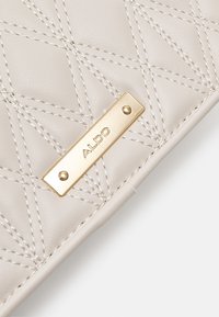 Gros plan sur cuir matelassé beige avec coutures beiges et plaque métallique dorée embossée du nom de la marque ALDO.