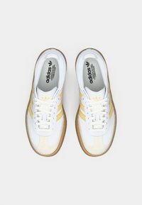 Białe trampki z żółtym wzorem trzech pasków, teksturowaną zamszową nakładką na palce oraz gumową podeszwą. Posiadają zapięcie na sznurówki oraz wkładkę z logo.