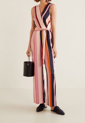 Kobieta w bezrękawowym, wielokolorowym kombinezonie w pionowe paski z paskiem, trzymająca czarną cylindryczną torebkę oraz nosząca czarne buty wsuwane.