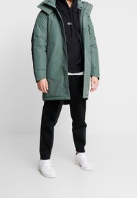 Grön parkas med dragkedja fram, justerbar huva och sidofickor, lagd över en svart hoodie; matchad med svarta byxor och vita sneakers.